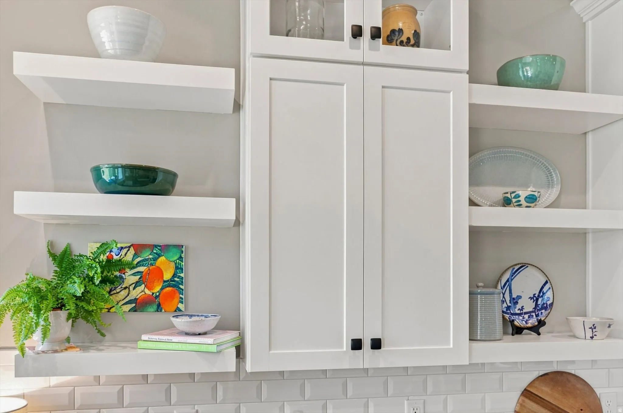 White shaker-style cabinetry with matte black hardware, floating display shelving, upper glass-front cabinets, and a subway tile backsplash