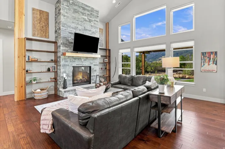 Vaulted ceiling living room featuring a stacked stone fireplace with wood mantel, hardwood flooring, built-in shelving, and large picture windows