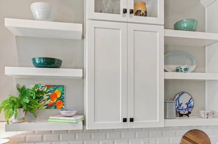 White shaker-style cabinetry with matte black hardware, floating display shelving, upper glass-front cabinets, and a subway tile backsplash