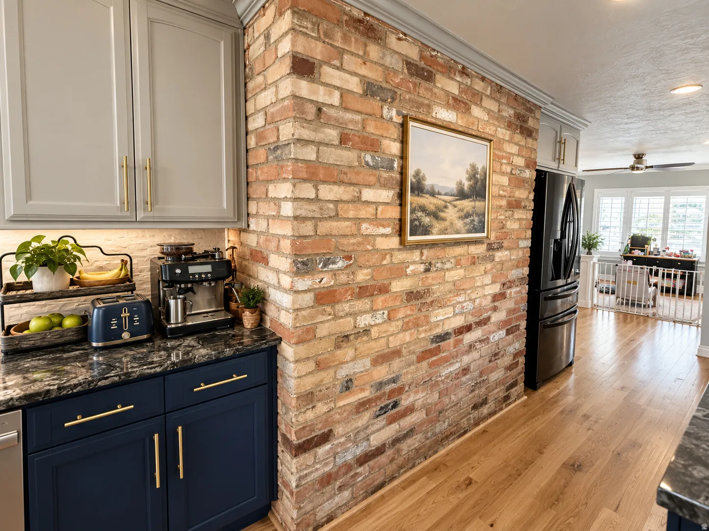 Exposed brick accent wall, wood-finish flooring, two-tone cabinetry with gold-tone hardware, dark countertop, and recessed lighting