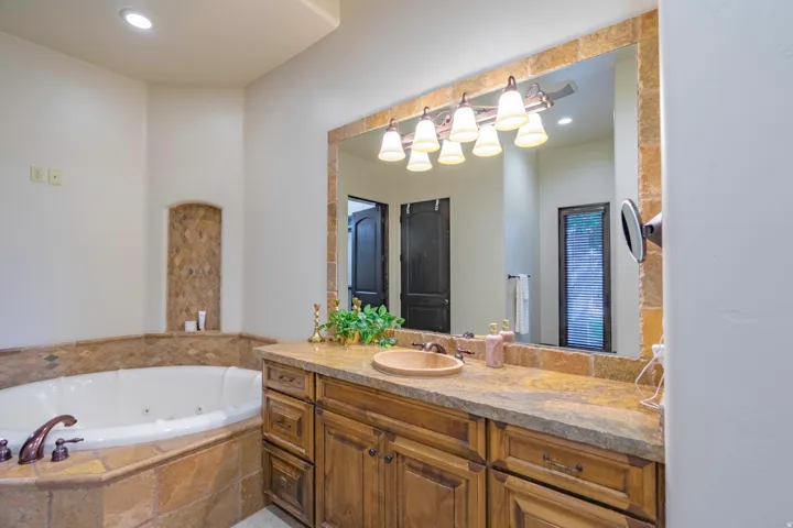 Jetted soaking tub with tile surround, wood-finish vanity, stone countertop with integrated sink, and large mirror with overhead lighting