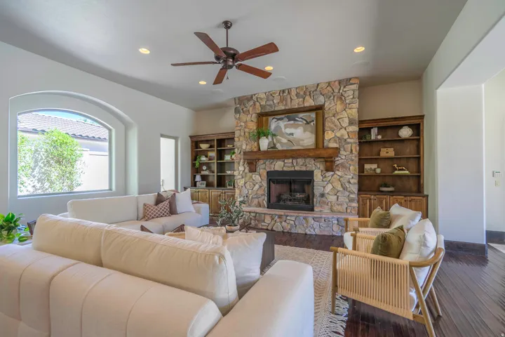 Stone-clad fireplace with a wood mantle, built-in shelving, arched window, ceiling fan, and recessed lighting