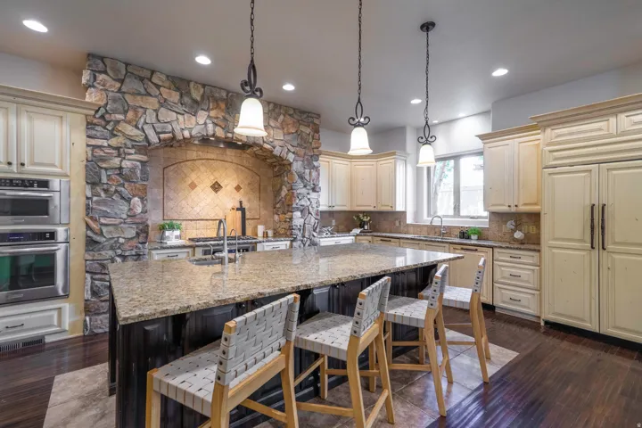 Stone-clad range hood with tile backsplash, stainless steel double ovens, large kitchen island with a granite countertop, pendant lighting, and cream-colored cabinetry