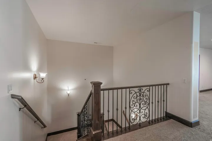 Carpeted flooring, decorative wrought iron banister, wood newel post, and wall-mounted sconce lighting