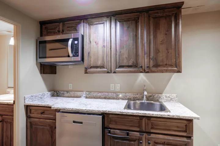 Stained wood cabinetry with integrated stainless steel microwave, stainless steel dishwasher, stainless steel sink, and light-toned stone countertops