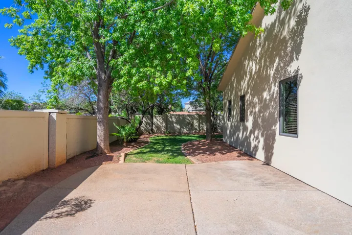 Private yard featuring a concrete patio, landscaped beds with mature trees, and a grass area enclosed by a perimeter wall