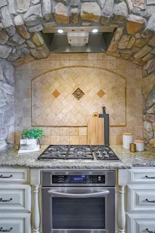 Stone archway detailing, stainless steel range hood, integrated gas cooktop, built-in stainless steel oven, and light-toned cabinetry
