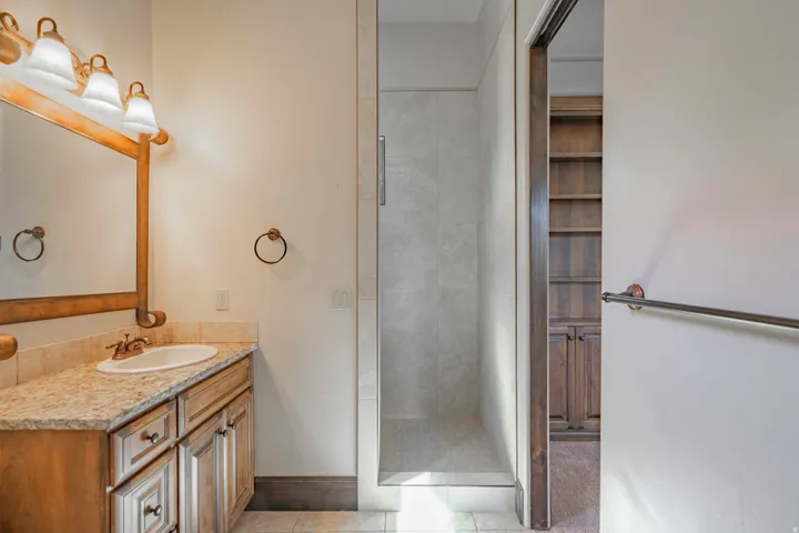 Granite-finish countertop with an undermount sink, wood-finish vanity cabinetry, wood-framed mirror, and a walk-in shower with tile surround