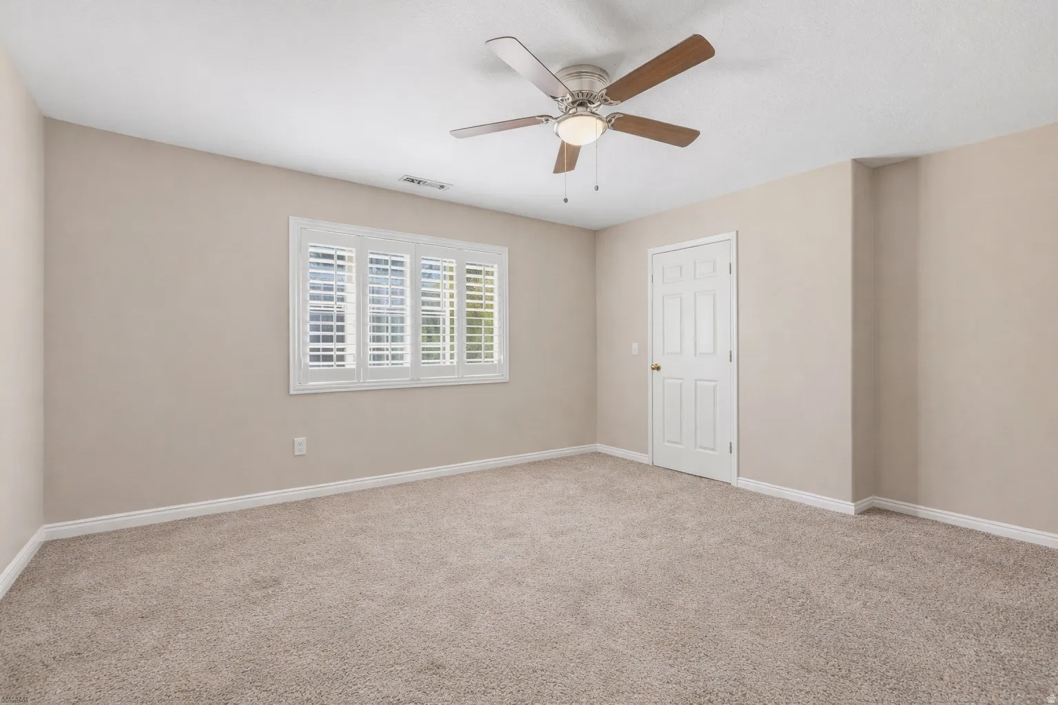 Expansive room featuring neutral-toned carpeting, a ceiling fan with integrated lighting, a multi-pane window with white plantation shutters, and a paneled interior door