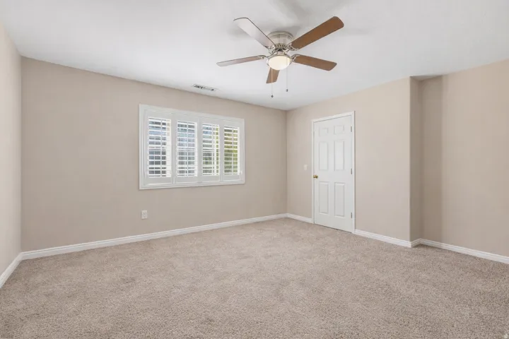 Expansive room featuring neutral-toned carpeting, a ceiling fan with integrated lighting, a multi-pane window with white plantation shutters, and a paneled interior door