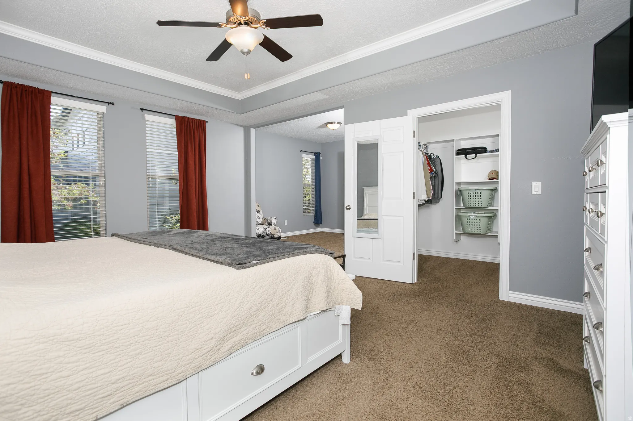 Tray ceiling with crown molding, ceiling fan with integrated lighting, two windows with blinds, built-in closet shelving, and a mirrored closet door