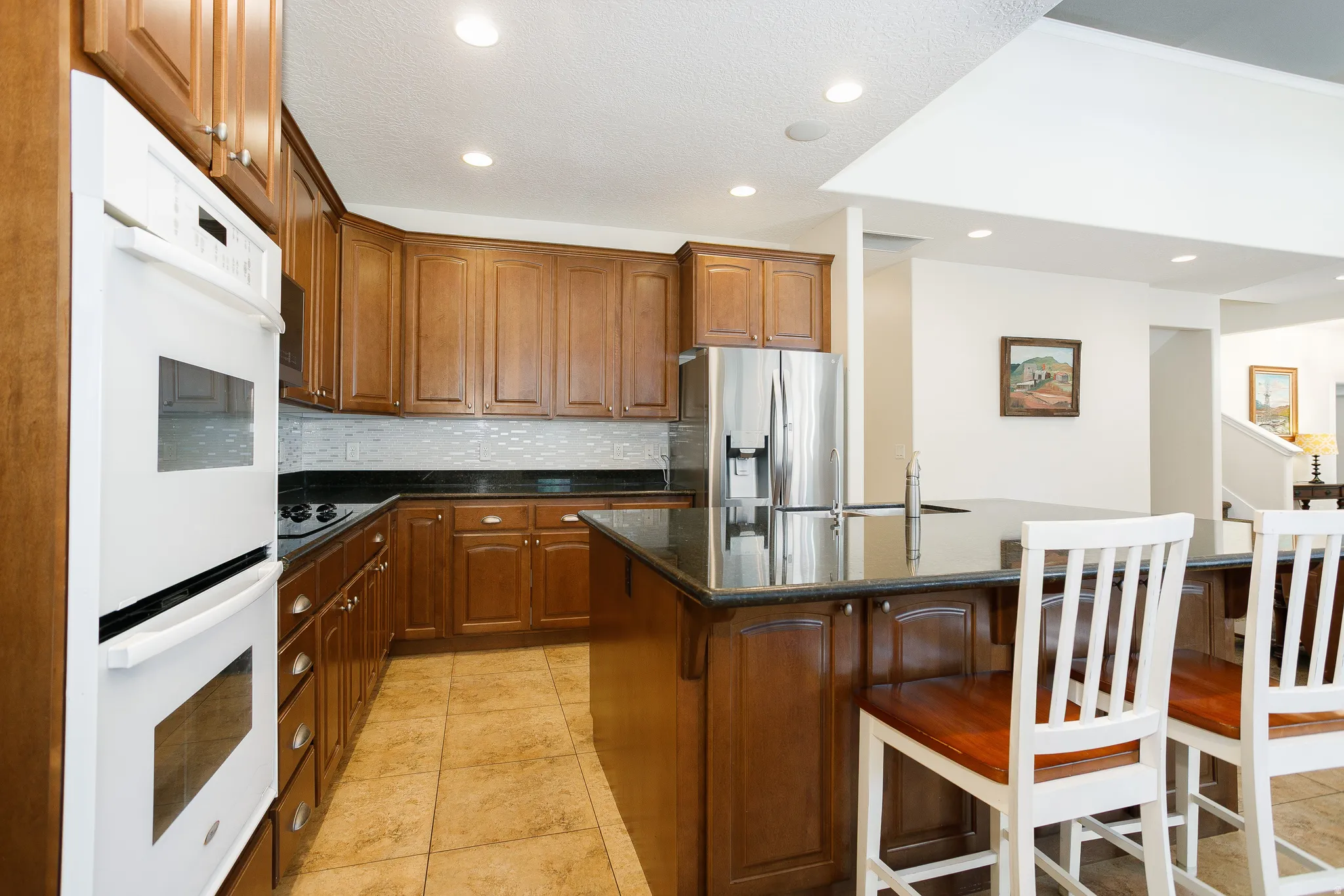 Built-in double ovens, stainless steel refrigerator, dark countertops, tiled backsplash, and recessed lighting