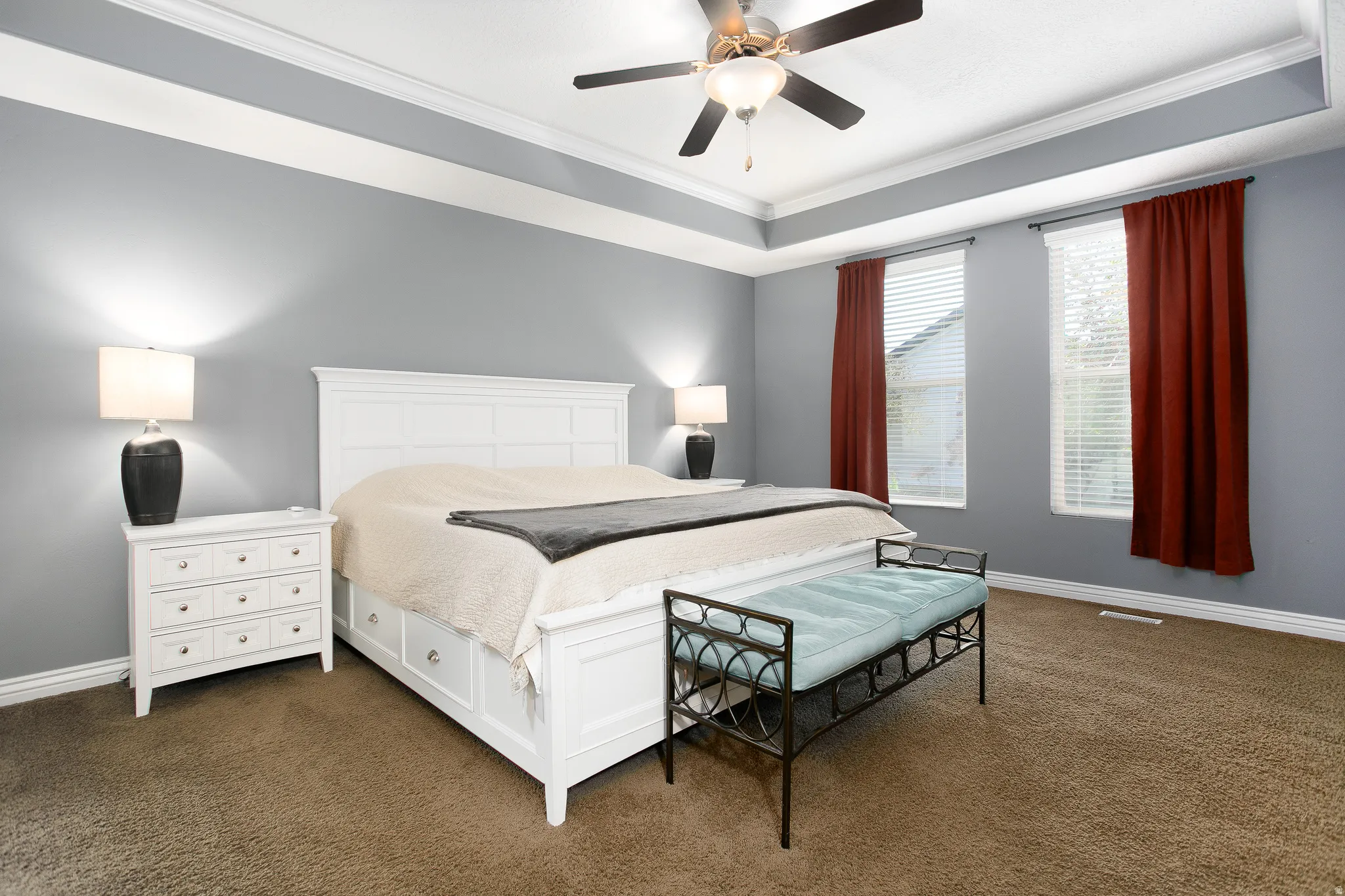 Tray ceiling with crown molding, ceiling fan, two windows with blinds, and carpeted flooring