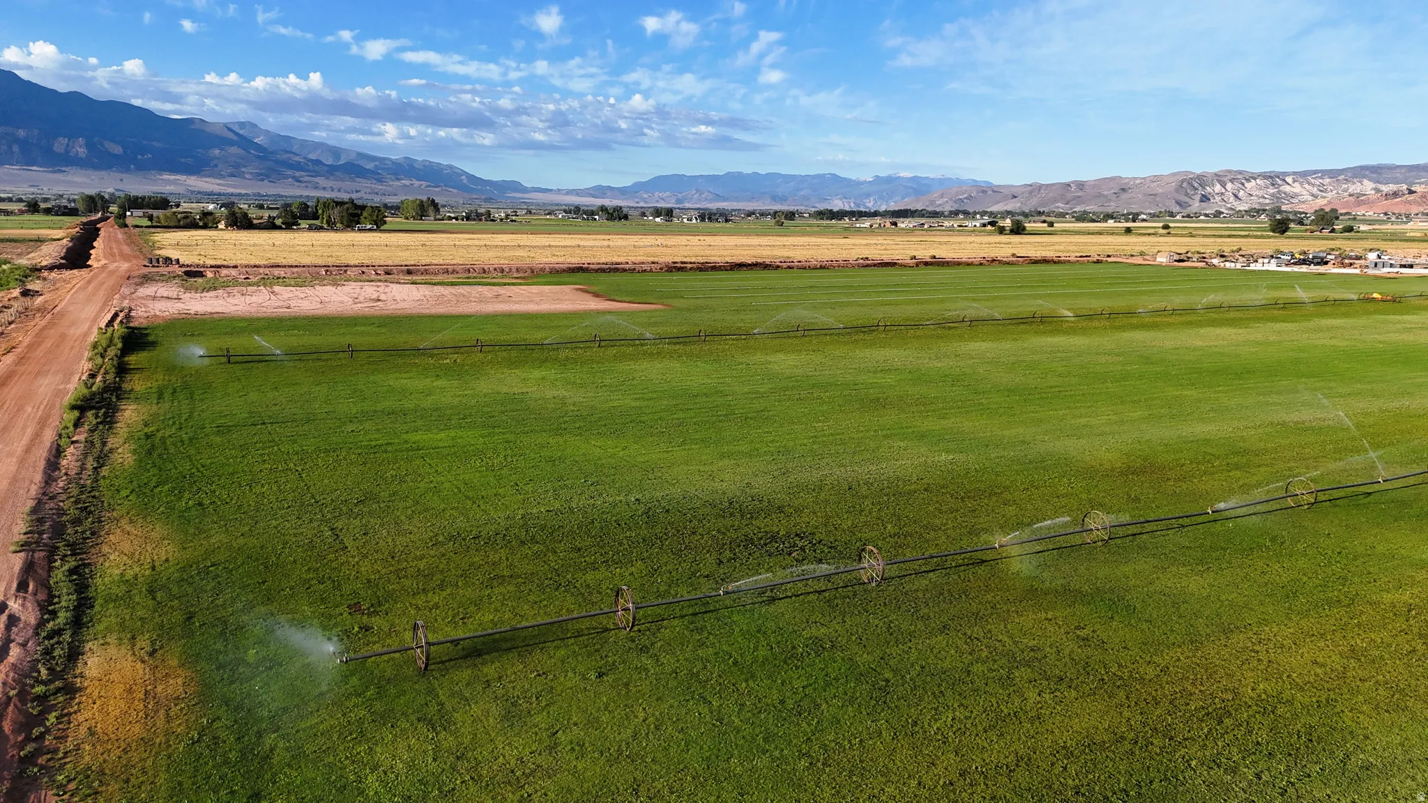 Expansive green fields with active irrigation systems, bordered by a dirt road, and backed by distant mountains