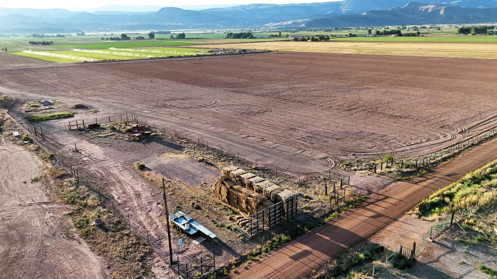 Expansive agricultural land featuring a dirt road, partial fencing, and distant mountain scenery