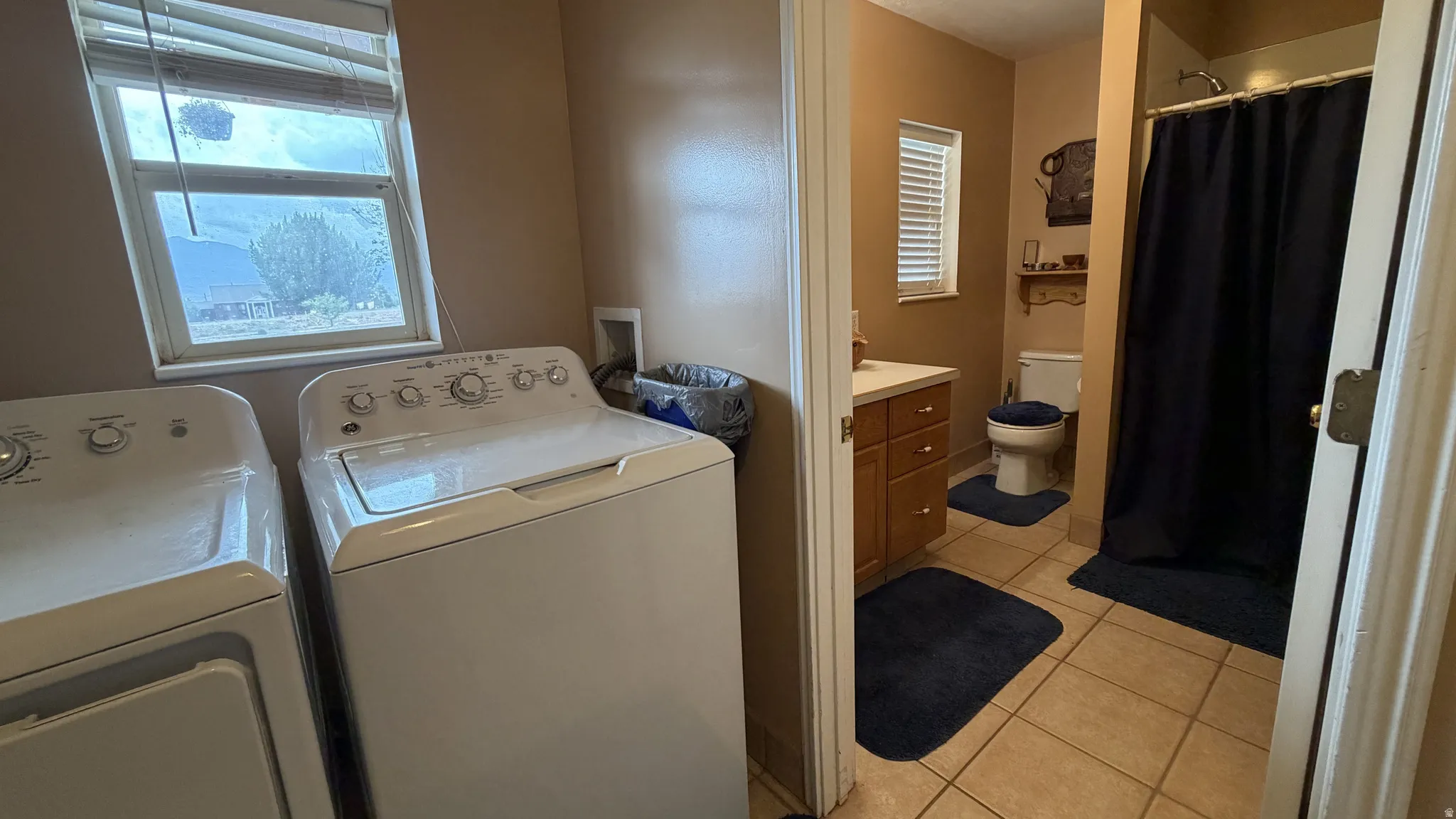 Laundry area with a window and nearby bathroom featuring a vanity with a light countertop, wood-finish cabinetry, and a shower with a tiled surround