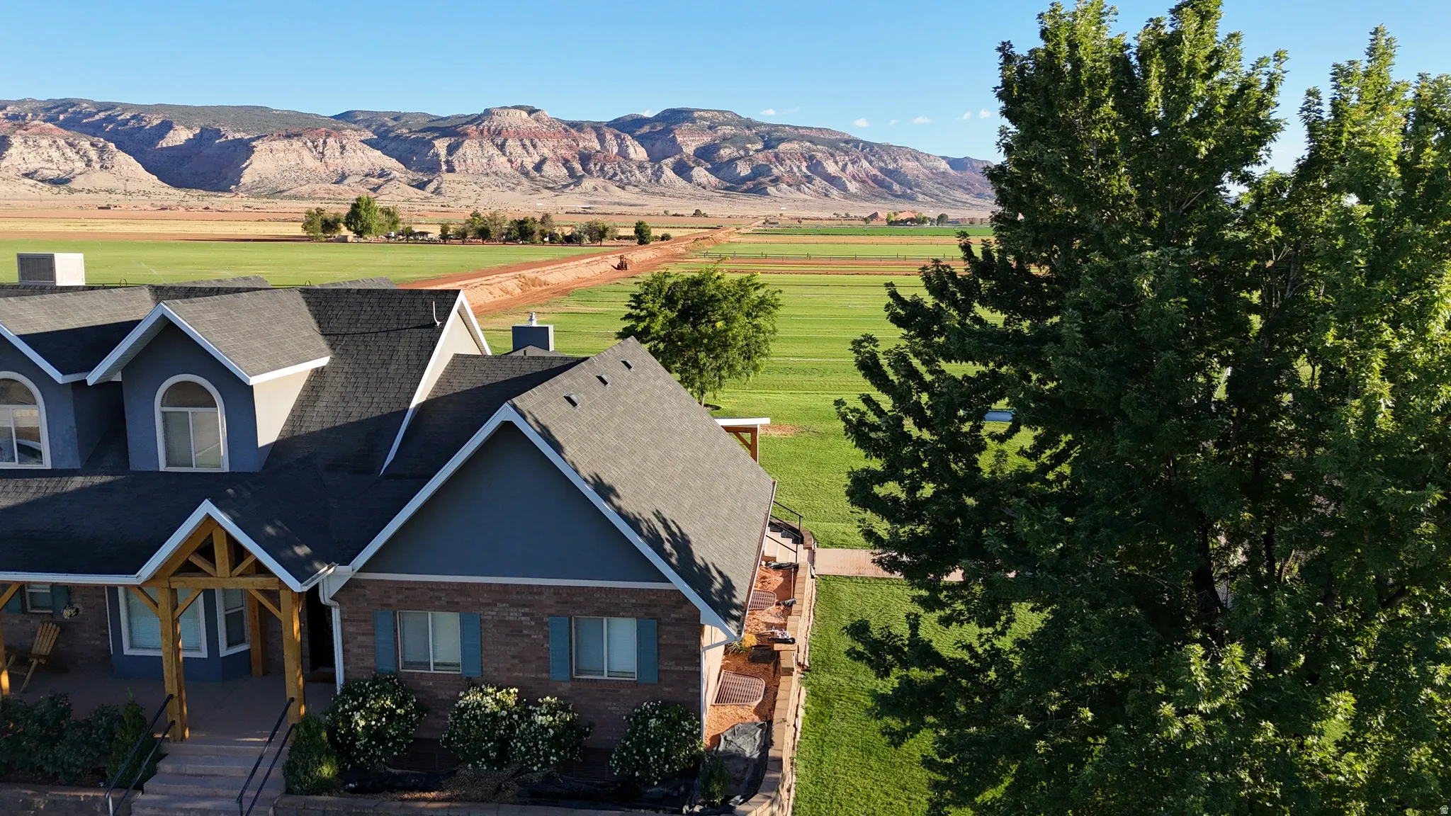 Gable roofline with dormer windows, brick and siding exterior, front porch with wood beam accents, expansive green fields, and distant mountain range
