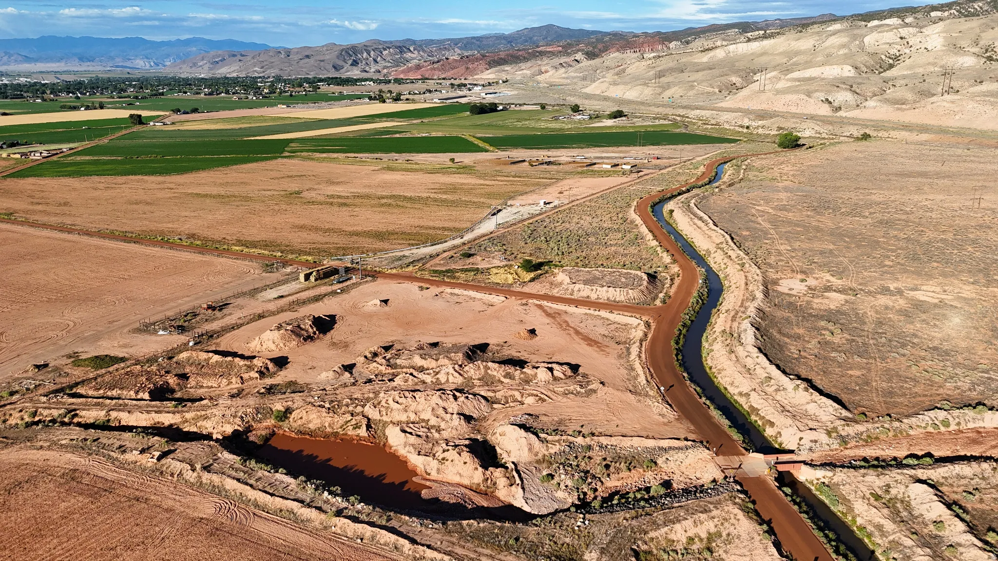 Expansive undeveloped land featuring a meandering water canal, a dirt access road, and distant agricultural fields