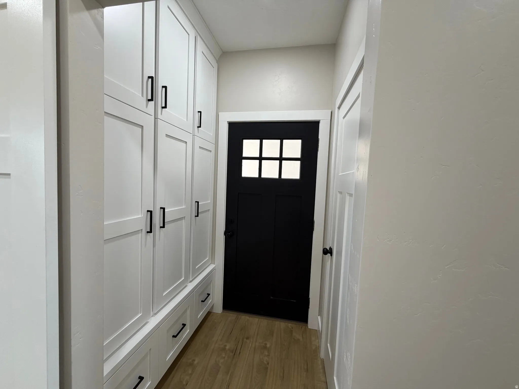 Built-in white shaker lockers with black hardware, wood-finish flooring, and a black exterior door with window panes going into the garage.