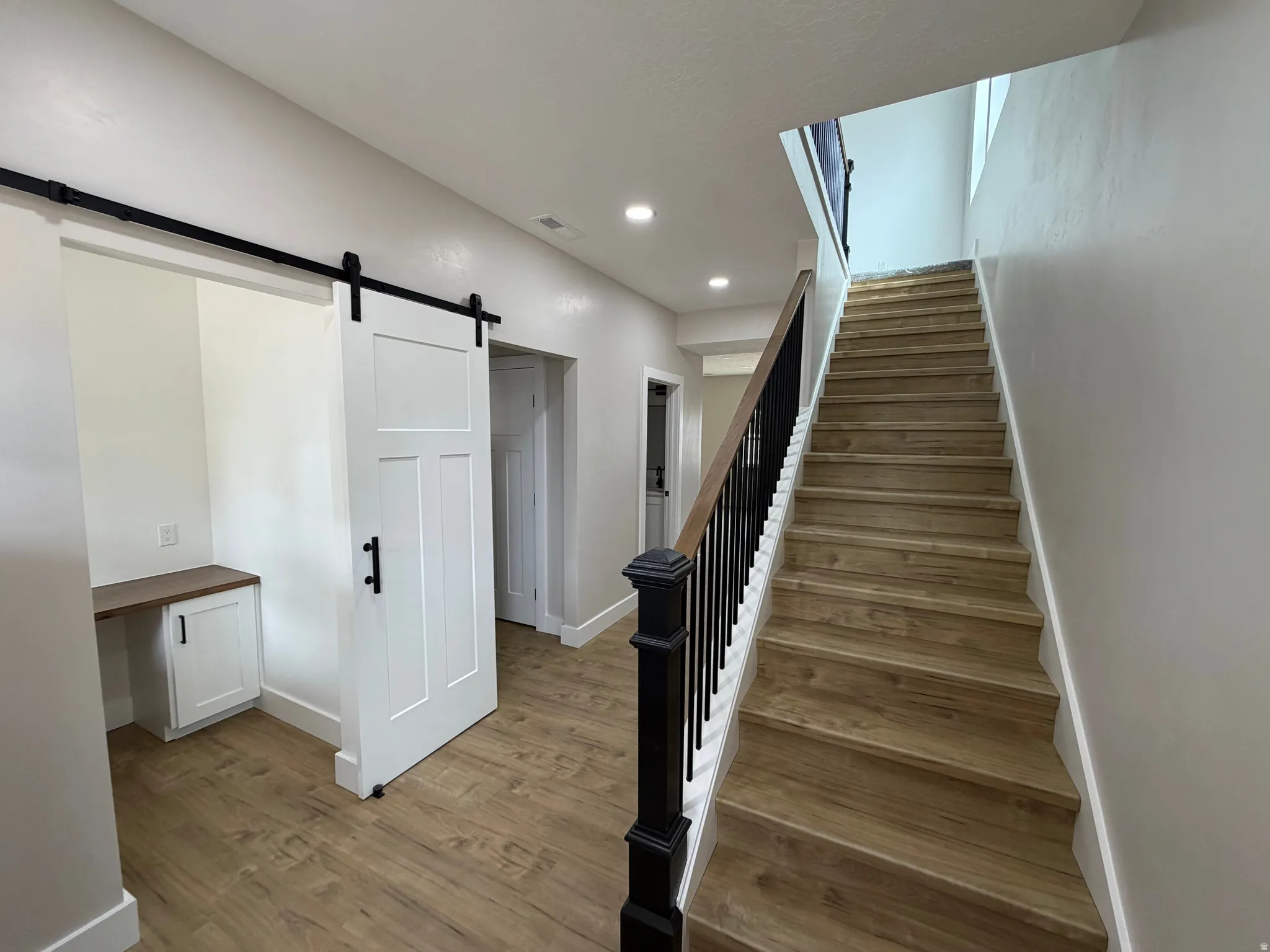 Wood-finish flooring, white walls, built-in desk, barn door, and a staircase with black balusters