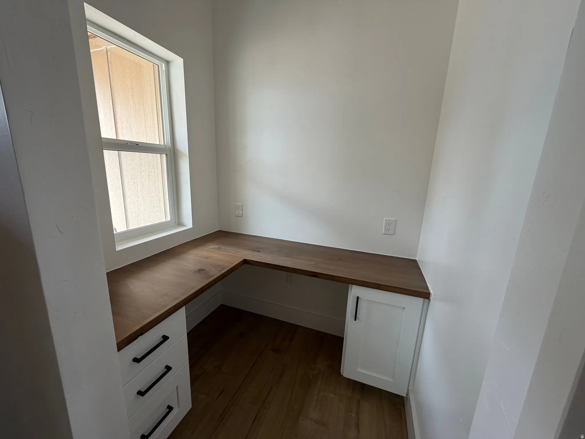 Built-in L-shaped desk with wood-finish countertop, white cabinetry with black hardware, and wood-finish flooring