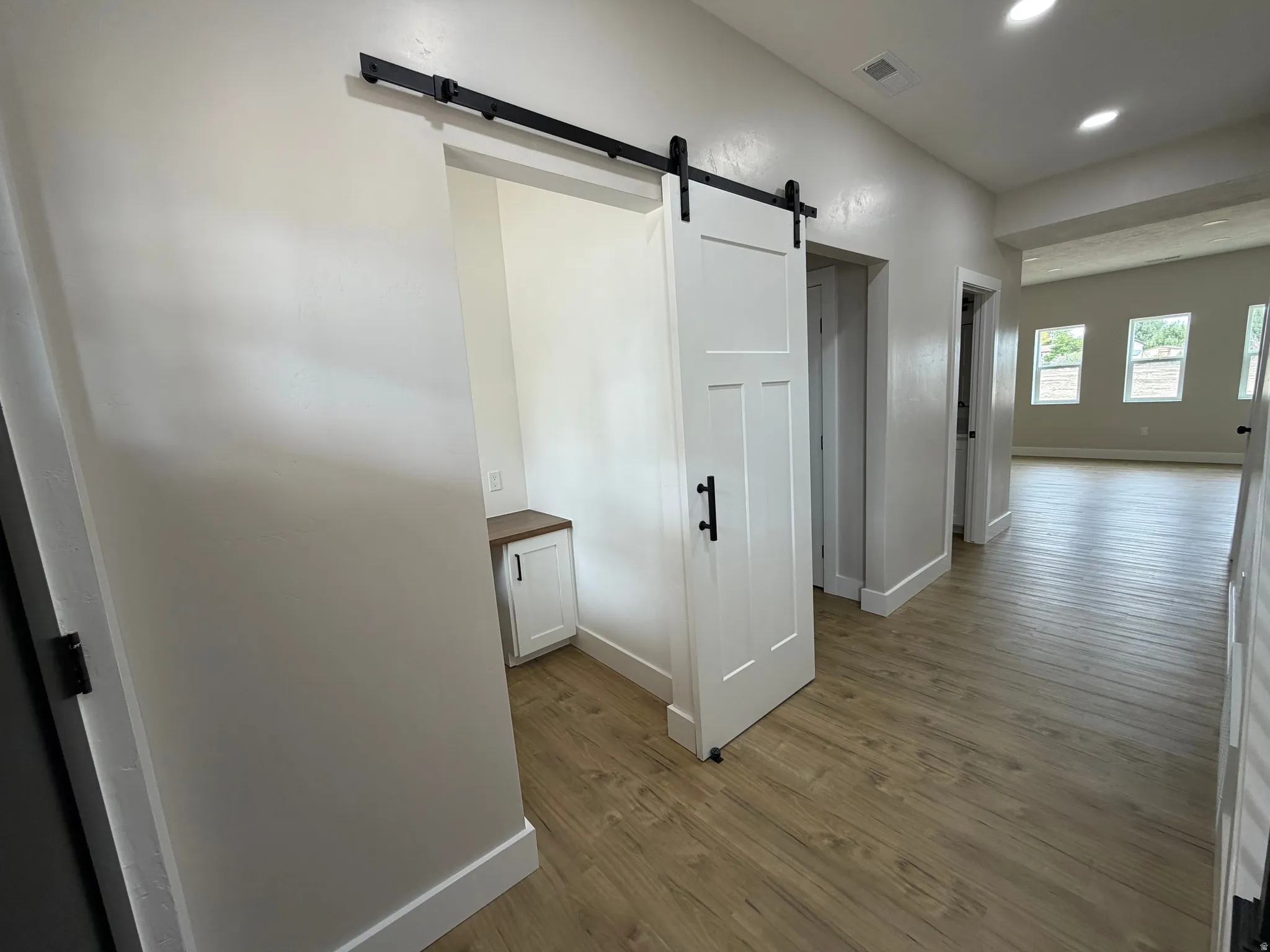 White barn door with black hardware, wood-finish flooring, recessed lighting, built-in desk with wood-finish countertop, and white cabinetry
