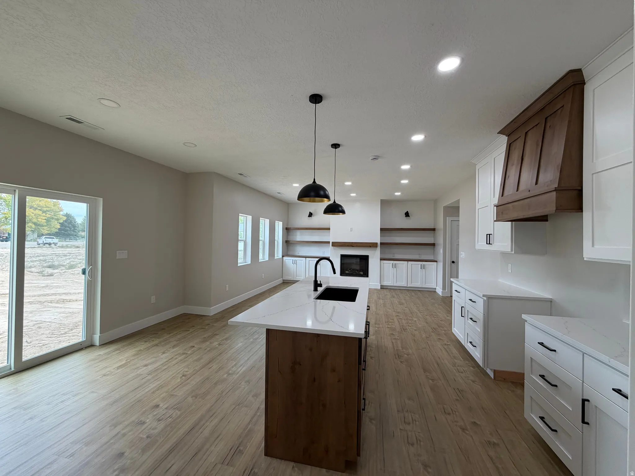 Open-concept kitchen and living area featuring wood-finish flooring, white cabinetry with matte black hardware, a kitchen island with a white countertop, a recessed fireplace, and a wood range hood