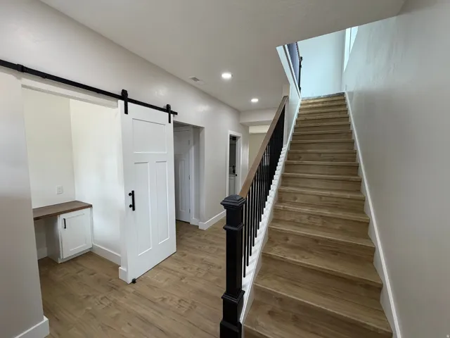 Wood-finish flooring, white walls, built-in desk, barn door, and a staircase with black balusters