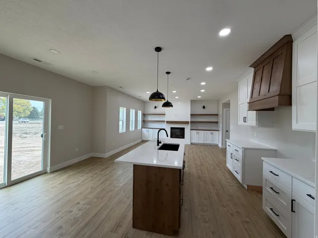 Open-concept kitchen and living area featuring wood-finish flooring, white cabinetry with matte black hardware, a kitchen island with a white countertop, a recessed fireplace, and a wood range hood