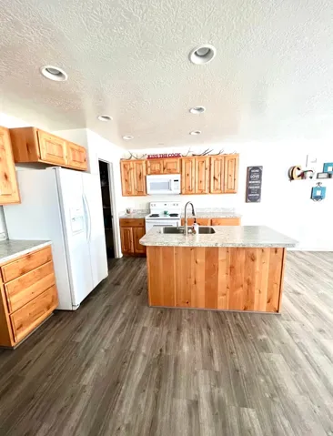 Kitchen featuring wood-finish cabinetry, light-toned countertops, a central island with an integrated sink, recessed ceiling lighting, and wood-finish flooring