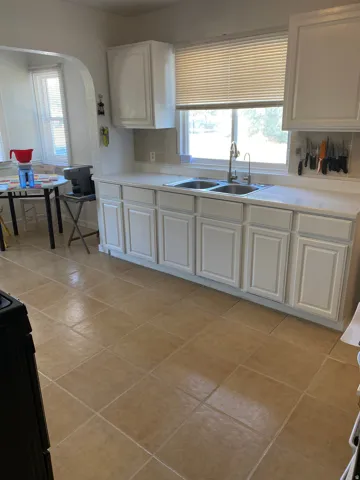 White cabinetry with brushed nickel hardware, dual basin stainless steel sink, light-toned countertops, and square tile flooring