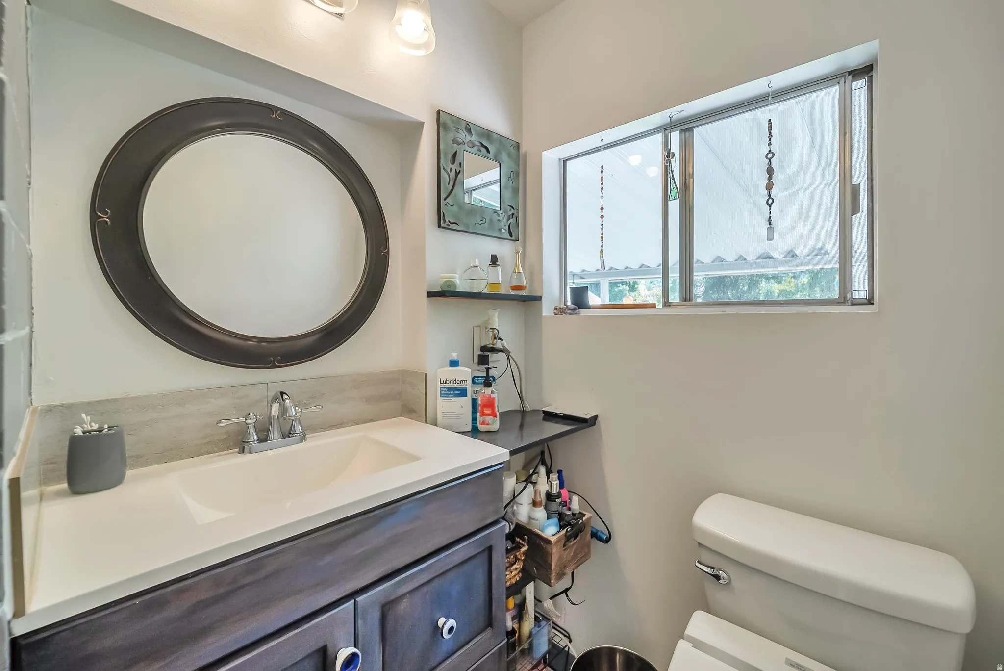 Integrated sink with a light-toned countertop, wood-finish vanity with cabinet storage, round wall-mounted mirror, and a fixed window