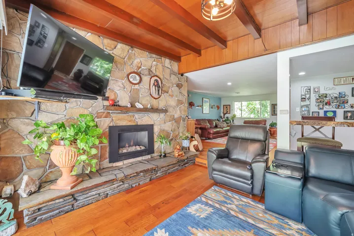 Stone-clad fireplace with a gas insert, wood-finish flooring, exposed wood beam ceiling, and a pass-through opening to an additional room