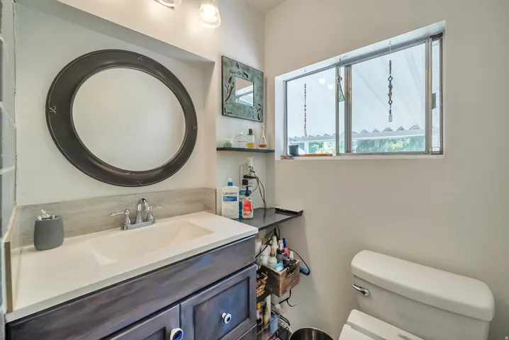 Integrated sink with a light-toned countertop, wood-finish vanity with cabinet storage, round wall-mounted mirror, and a fixed window