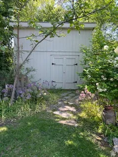 White paneled shed with double doors featuring black hardware, surrounded by lush green foliage and flowering plants