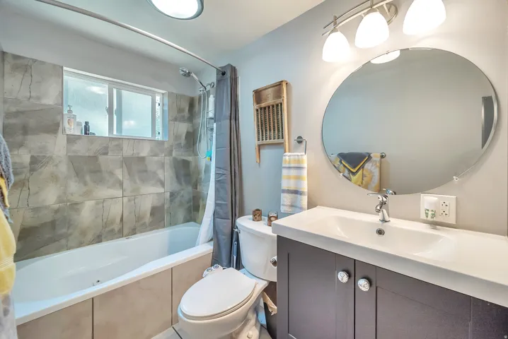 Bathroom featuring a single vanity with storage, an oval mirror, a three-light fixture, and a tiled tub surround with a window