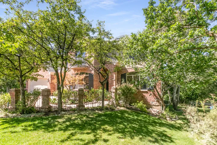 Brick exterior residence with upper-level balcony, ground-level windows, and a landscaped front yard featuring mature trees and a decorative fence
