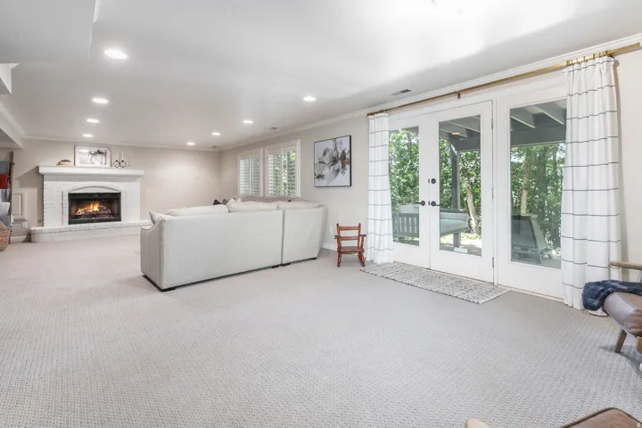 Recessed lighting, brick fireplace with a white mantel, white double French doors, large windows with shutters, and light-toned carpeting