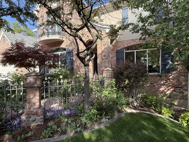 Brick facade with stone accents, bay window with dark shutters, wrought iron balcony, mature landscaping, and decorative metal fencing