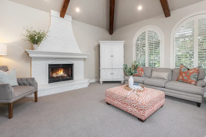 Vaulted ceiling with exposed wood beams, white brick fireplace, arched windows with plantation shutters, and light-toned carpeting