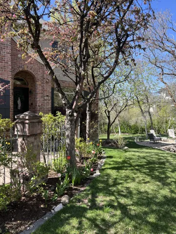 Brick exterior with arched entry, mature trees, manicured lawn, and planted flower beds