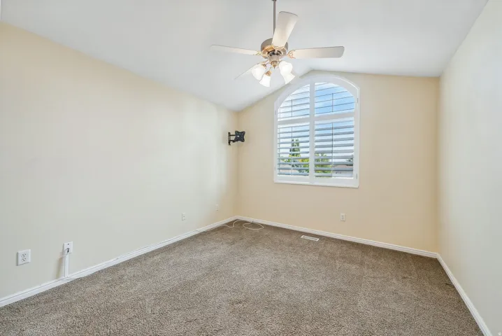 Vaulted ceiling with a ceiling fan, arched window with plantation shutters, wall-mounted television bracket, and light-toned carpeting