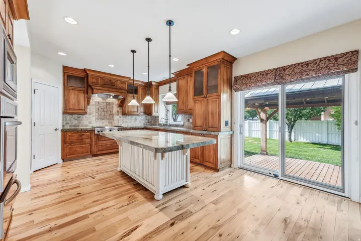 Wood-finish flooring throughout, kitchen with wood cabinetry and built-in appliances, central island with seating, pendant lighting, and sliding glass door to outdoor decking