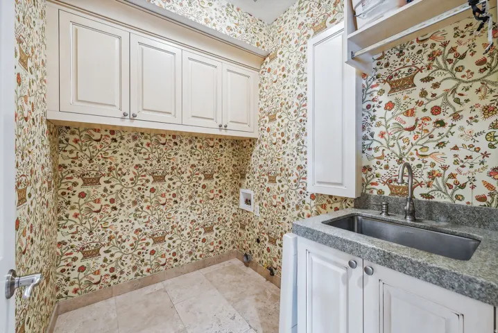 Laundry area featuring light-toned cabinetry, a utility sink with a stone-finish countertop, and decorative wall covering