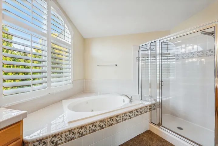 Soaking tub with jetted features, glass-enclosed shower, arched window with plantation shutters, decorative tile accents, and tiled tub surround