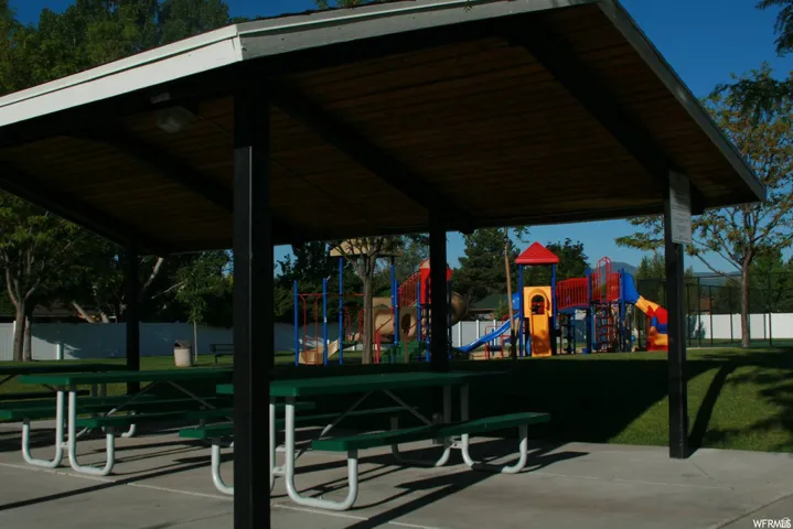 Covered patio structure with concrete flooring and multiple picnic tables, overlooking a playground area