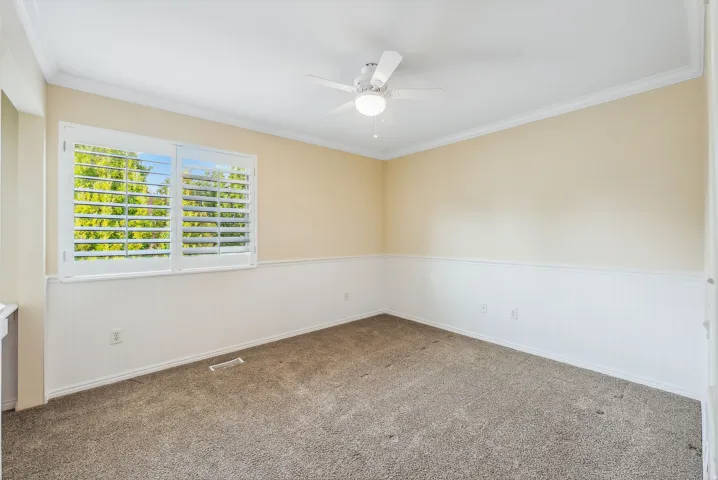 Carpeted room featuring a ceiling fan with integrated lighting, crown molding, white plantation shutters, and wainscoting