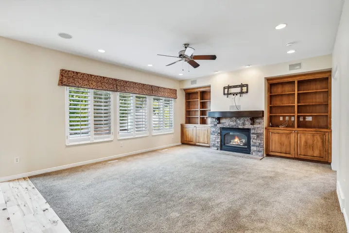 Built-in shelving with lower cabinetry, stone-surround fireplace with a wood mantel, multiple windows with plantation shutters, recessed lighting, and a ceiling fan