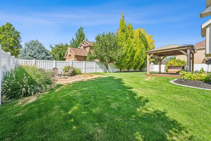 Expansive green lawn with a white privacy fence, a built-in gazebo on a raised deck, a stone fire pit area, and established perimeter landscaping