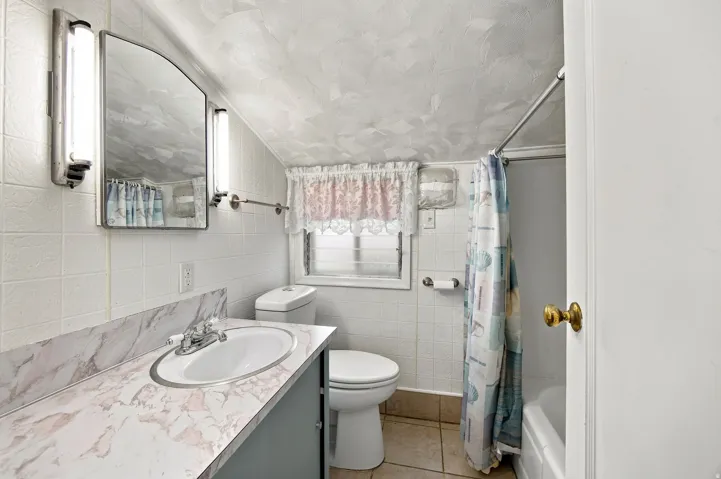 White tile walls, vanity with a white countertop featuring an integrated sink, dual wall-mounted light fixtures, built-in medicine cabinet, and a window